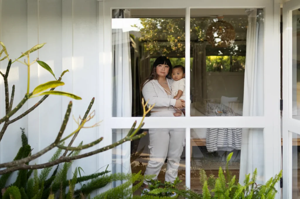 Mother holding baby standing at a bright sliding glass door surrounded by greenery